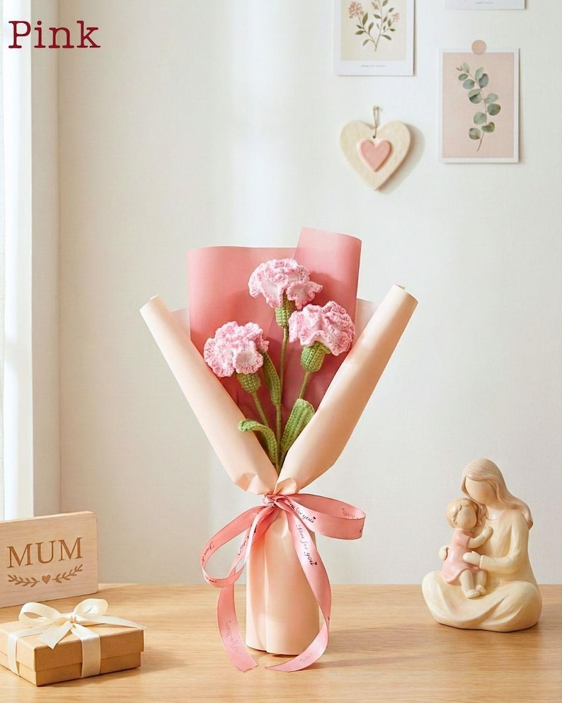 May include: A bouquet of pink crocheted carnations wrapped in pink paper and tied with a pink ribbon. The word "Pink" is in the top left corner. A small gift box and a wooden sign that says "MUM" are also visible.