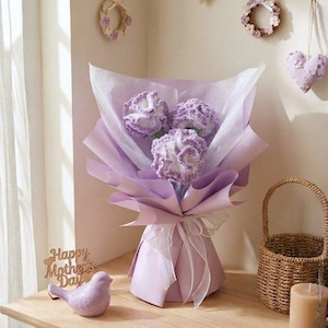 May include: A bouquet of three crocheted lavender flowers wrapped in purple paper and tied with a white ribbon. A small ceramic bird and a wooden sign that says "Happy Mother's Day" are on a wooden table.