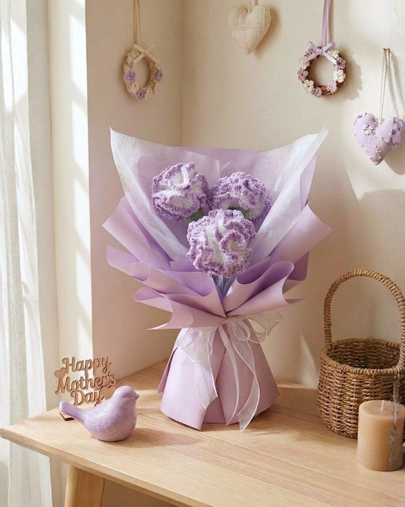 May include: A bouquet of three crocheted lavender flowers wrapped in purple paper and tied with a white ribbon. A small ceramic bird and a wooden sign that says "Happy Mother's Day" are on a wooden table.