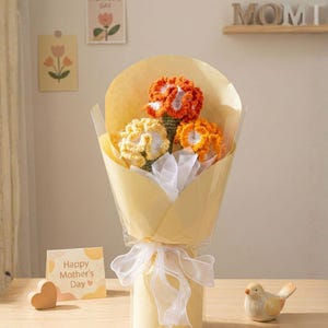 May include: A bouquet of crocheted flowers in shades of orange and yellow, wrapped in yellow paper and tied with a white ribbon. A small card with "Happy Mother's Day" is visible. A wooden heart and a ceramic bird are also present.