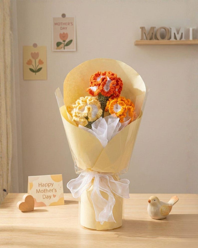May include: A bouquet of crocheted flowers in shades of orange and yellow, wrapped in yellow paper and tied with a white ribbon. A small card with "Happy Mother's Day" is visible. A wooden heart and a ceramic bird are also present.