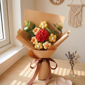 May include: A bouquet of handmade crochet flowers wrapped in brown paper and tied with a brown ribbon. The bouquet features yellow and orange flowers with a central red flower, and green leaves. A small heart-shaped dish and a vase of dried flowers are also visible.