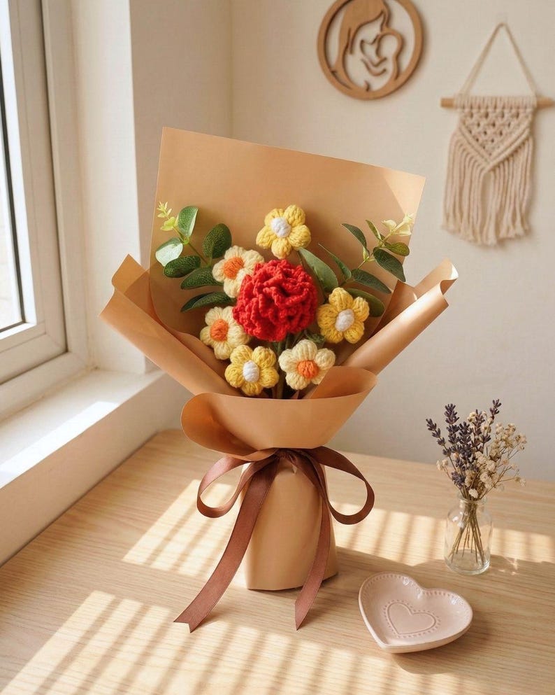 May include: A bouquet of handmade crochet flowers wrapped in brown paper and tied with a brown ribbon. The bouquet features yellow and orange flowers with a central red flower, and green leaves. A small heart-shaped dish and a vase of dried flowers are also visible.
