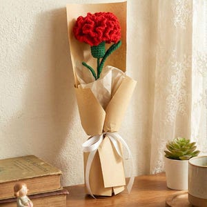 May include: A single red crocheted carnation flower wrapped in brown paper, tied with a white ribbon. The flower has a green stem and is presented as a gift. The background includes a wooden surface and a neutral wall.
