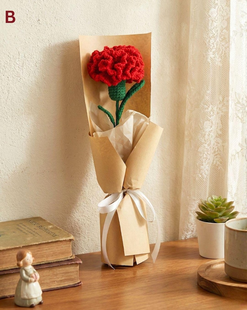 May include: A single red crocheted carnation flower wrapped in brown paper, tied with a white ribbon. The flower has a green stem and is presented as a gift. The background includes a wooden surface and a neutral wall.