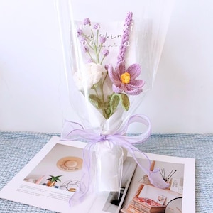 K&ouml;nnte beinhalten: Ein Strau&szlig; geh&auml;kelter Blumen in Lila- und Wei&szlig;t&ouml;nen, verpackt in durchsichtige Cellophanfolie und mit einem Lavendelband gebunden. Der Strau&szlig; liegt auf einem Buch.