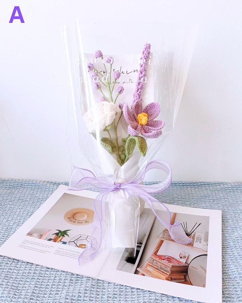 K&ouml;nnte beinhalten: Ein Strau&szlig; geh&auml;kelter Blumen in Lila- und Wei&szlig;t&ouml;nen, verpackt in durchsichtige Cellophanfolie und mit einem Lavendelband gebunden. Der Strau&szlig; liegt auf einem Buch.