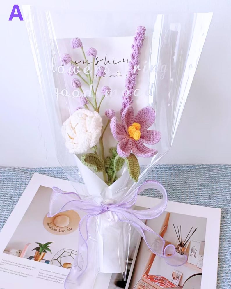 K&ouml;nnte beinhalten: Ein Strau&szlig; geh&auml;kelter Blumen in Lila- und Wei&szlig;t&ouml;nen, verpackt in durchsichtige Cellophanfolie und mit einem Lavendelband gebunden. Der Strau&szlig; liegt auf einem Buch mit dem sichtbaren Text "sunshine is with you".