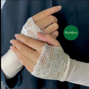 May include: Ivory lace fingerless gloves. The gloves feature a delicate floral pattern and are worn on the hands. The text "RoyalJava" is in a green circle in the background. The gloves are a light color and appear to be made of a soft material.