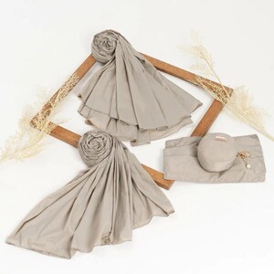 May include: A collection of beige scarves and a matching small round case with a rectangular pouch. The scarves are draped and rolled, and the case has a gold-coloured zip pull with a pearl charm. The items are arranged on a wooden frame.