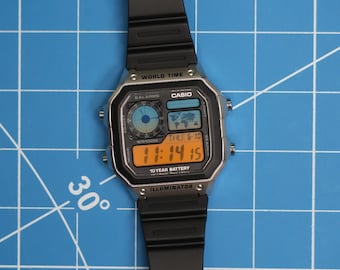 Puede incluir: Reloj de pulsera digital negro con esfera rectangular y correa negra. La esfera muestra la hora, la fecha y las funciones de hora mundial. El reloj tiene la marca "CASIO" y el texto "10 YEAR BATTERY". Sobre un fondo de cuadrícula azul.