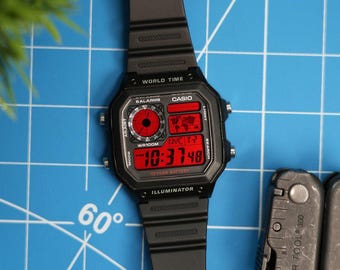 Puede incluir: Un reloj digital Casio negro con una pantalla roja. El reloj tiene una batería de 10 años y una función de iluminación. La esfera del reloj muestra la hora, la fecha y el día de la semana. El reloj está sobre un fondo de cuadrícula azul.