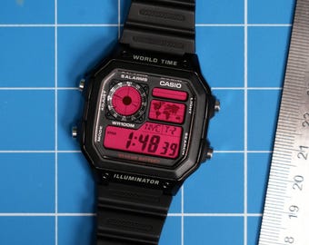 Puede incluir: Un reloj digital Casio negro con una pantalla roja. El reloj tiene una función de hora mundial, alarmas y una batería de 10 años. El reloj está sobre un fondo azul de cuadrícula.