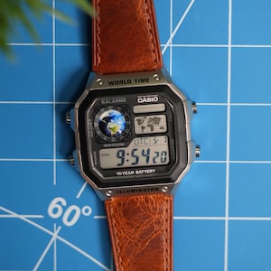 May include: A digital Casio watch with a brown leather band. The watch face displays the time, world time, and other functions. The watch is set to 9:54:20 PM. The watch has a silver-colored case and is on a blue surface.