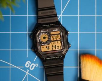 Puede incluir: Un reloj digital Casio negro con una esfera dorada y una correa negra. El reloj tiene una función de hora mundial, alarmas y un iluminador. El reloj está sobre un fondo azul de cuadrícula.
