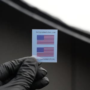 Puede incluir: Una pequeña pegatina rectangular con dos banderas americanas. Las banderas tienen rayas rojas y blancas y un campo azul con estrellas blancas. El texto "JellyLabWatches.com" está impreso en la pegatina.