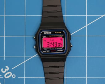 Puede incluir: Reloj digital Casio F-91W negro con correa negra. La esfera muestra la hora 3:49:05 PM, el día de la semana y la fecha. El reloj tiene un borde azul y el texto "CASIO F-91W".