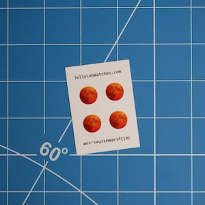 May include: A white card with four red moon stickers, against a blue grid background. The stickers have a textured surface. The card has the text "JellyLabWatches.com".