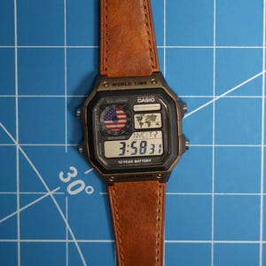 May include: A digital watch with a brown leather strap. The watch face displays the time, 3:58:31, and features a small American flag graphic. The watch also has the text "10 YEAR BATTERY".