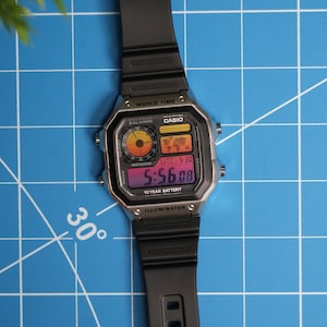 May include: A black digital Casio watch with a rectangular face and black strap. The watch displays the time 5:56:08, with world time and a 10-year battery. The watch is set against a blue grid background.