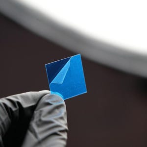 Puede incluir: Una película adhesiva azul cuadrada con una esquina despegada, sostenida por una mano enguantada. La película es de un color azul vibrante, y la esquina despegada revela un tono más claro. El fondo es un degradado borroso de blanco y marrón.