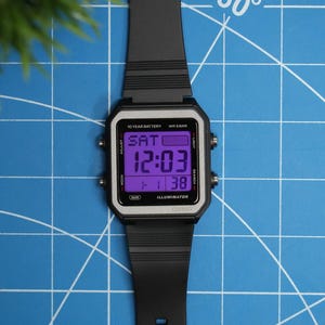 May include: A black digital wristwatch with a square silver-toned bezel and a purple backlit display. The screen shows the text SAT, 12:03, 1-1, 38, 10 YEAR BATTERY, WR 5BAR, and ILLUMINATOR. It features a black resin strap and four side buttons.