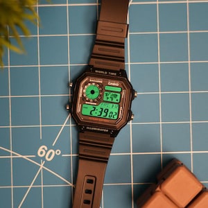 Può includere: Un orologio da polso digitale nero con un display verde. Il quadrante dell'orologio mostra l'ora, la data e il giorno della settimana. L'orologio ha un cinturino nero e una cassa nera. L'orologio è su uno sfondo a griglia blu e bianco.