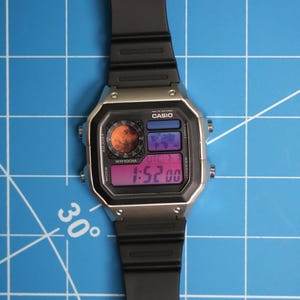 May include: A digital Casio watch with a black strap and silver case. The watch face shows the time, date, and other functions. The watch is on a blue grid background. The time displayed is 1:52.