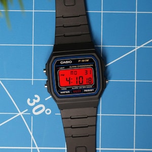 May include: A black Casio F-91W digital watch with a red display showing the time 4:10:18. The watch has a black strap and a blue bezel. The watch is water resistant and has an alarm chronograph.