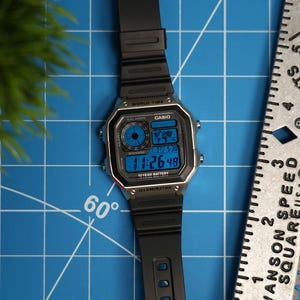 Op de afbeelding: Een zwarte Casio digitale horloge met een zilveren rand en een zwarte band. De wijzerplaat toont de tijd in blauwe cijfers. Het horloge ligt op een blauw rasteroppervlak naast een liniaal.
