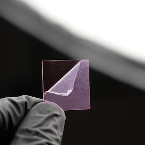 May include: A square, translucent, pinkish-purple object with a corner of the top layer peeled back, revealing a lighter layer underneath. The object is held by a gloved hand against a dark background.