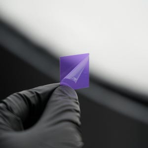 Puede incluir: Una pequeña película cuadrada de color púrpura se está despegando de una hoja de respaldo. La película está siendo sostenida por una mano enguantada.