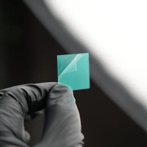 Op de afbeelding: Een vierkant, turquoise object met een gedeeltelijk afgetrokken toplaag wordt vastgehouden door een gehandschoende hand. Het object lijkt een dun, doorschijnend materiaal te zijn, mogelijk een sticker of film. De achtergrond is wazig, met een helder wit gebied.
