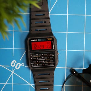 May include: Black Casio digital watch with a red digital display showing the time. The watch has a rectangular face with buttons and a black strap. The watch is water resistant and has alarm and chrono functions.