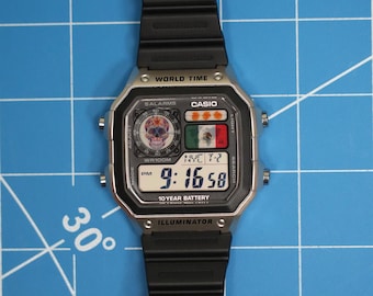 Puede incluir: Reloj digital Casio con correa negra y caja plateada. La esfera muestra la hora, un gráfico de calavera y la bandera mexicana. Funciones: hora mundial, alarmas y batería de 10 años.