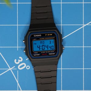 May include: Black Casio F-91W digital watch with a blue digital display showing the time. The watch has a black strap and is set against a blue grid background. The watch face displays the time as 4:04:40.