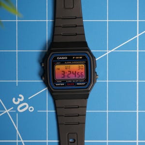 May include: A black Casio F-91W digital watch with a blue and red display. The watch has a black resin band and a water-resistant design. The display shows the time, date, and day of the week.