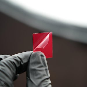 May include: A red adhesive sticker with a clear backing, partially peeled back to reveal the adhesive.
