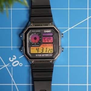 May include: A black Casio digital watch with a silver face and a black rubber strap. The watch has a world time function and a 10-year battery. The watch face displays the time, date, and day of the week. The watch is on a blue grid background.
