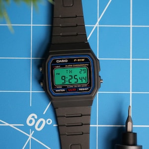 Könnte beinhalten: Eine schwarze Casio F-91W Digitaluhr mit blauem und rotem Rand. Das Zifferblatt der Uhr zeigt die Zeit, das Datum und den Wochentag an. Die Uhr befindet sich auf einem blauen Gitterhintergrund.