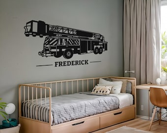 Könnte beinhalten: Schwarzes Wandbild eines Feuerwehrautos mit dem Namen "FREDERICK" darunter. Das Feuerwehrauto hat eine Leiter und ist an einer grauen Wand über einem Bett mit hellem Holzrahmen und Schubladen angebracht.