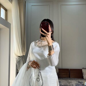 K&ouml;nnte beinhalten: Ein wei&szlig;es, lang&auml;rmeliges Kleid mit einem silbernen Perlenbesatz am Ausschnitt und einem passenden, durchsichtigen Dupatta. Das Kleid hat ein tailliertes Oberteil und einen flie&szlig;enden Rock. Das Model h&auml;lt eine silberne Clutch.