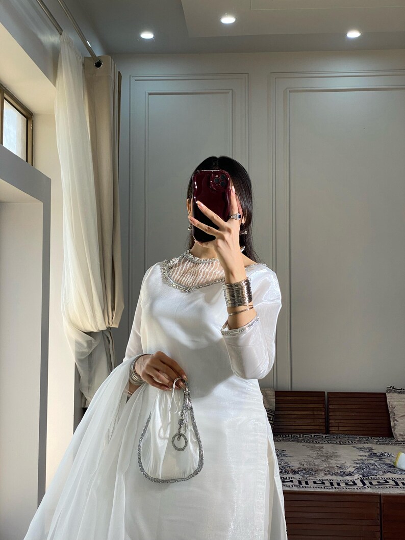 K&ouml;nnte beinhalten: Ein wei&szlig;es, lang&auml;rmeliges Kleid mit einem silbernen Perlenbesatz am Ausschnitt und einem passenden, durchsichtigen Dupatta. Das Kleid hat ein tailliertes Oberteil und einen flie&szlig;enden Rock. Das Model h&auml;lt eine silberne Clutch.