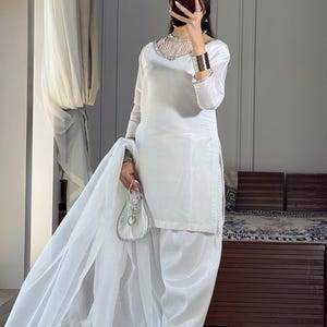 K&ouml;nnte beinhalten: Ein wei&szlig;es Salwar Kameez mit einem besetzten Ausschnitt und einem durchsichtigen Dupatta. Das Outfit wird mit wei&szlig;en Sandalen getragen.