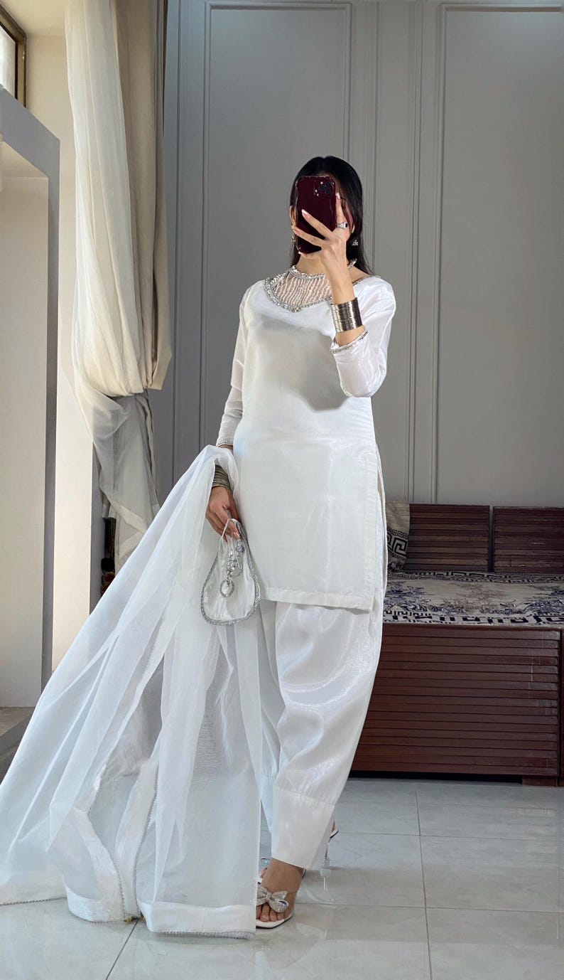 K&ouml;nnte beinhalten: Ein wei&szlig;es Salwar Kameez mit einem besetzten Ausschnitt und einem durchsichtigen Dupatta. Das Outfit wird mit wei&szlig;en Sandalen getragen.