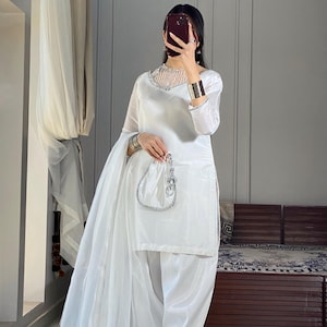 K&ouml;nnte beinhalten: Ein wei&szlig;es Salwar Kameez mit einem silbernen bestickten Ausschnitt und einem passenden Dupatta. Das Outfit wird mit einer wei&szlig;en Hose und silbernen Sandalen kombiniert.