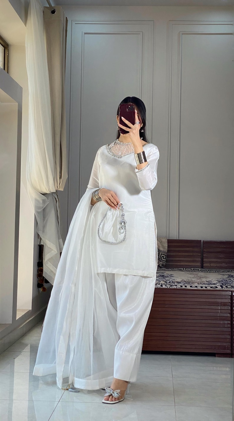 K&ouml;nnte beinhalten: Ein wei&szlig;es Salwar Kameez mit einem silbernen bestickten Ausschnitt und einem passenden Dupatta. Das Outfit wird mit einer wei&szlig;en Hose und silbernen Sandalen kombiniert.