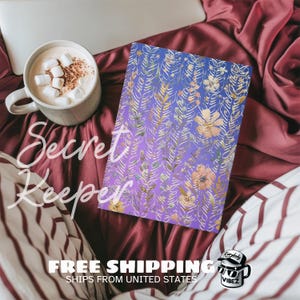 May include: A journal with a floral design in shades of purple, gold, and peach, with the words "Secret Keeper" in white script. A mug of hot chocolate with marshmallows is in the background.