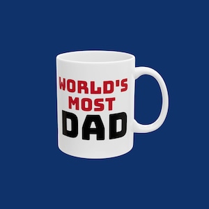 May include: White ceramic mug with the text "WORLD'S MOST DAD" in red and black block letters. The mug has a curved handle and is set against a dark blue background. A gift for Father's Day.