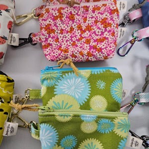 May include: Several small, zippered pouches with floral and patterned designs. The pouches have gold-colored clasps and are in shades of pink, green, and blue. Each pouch has a small tag attached. The pouches are likely for carrying small items.